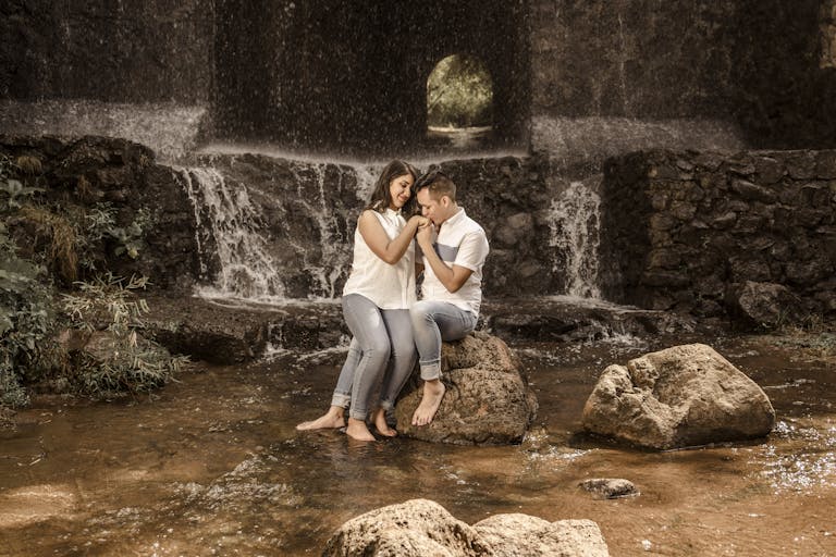 Juntos Seguros ICE A loving couple sits on rocks near a waterfall, embracing in a natural setting.