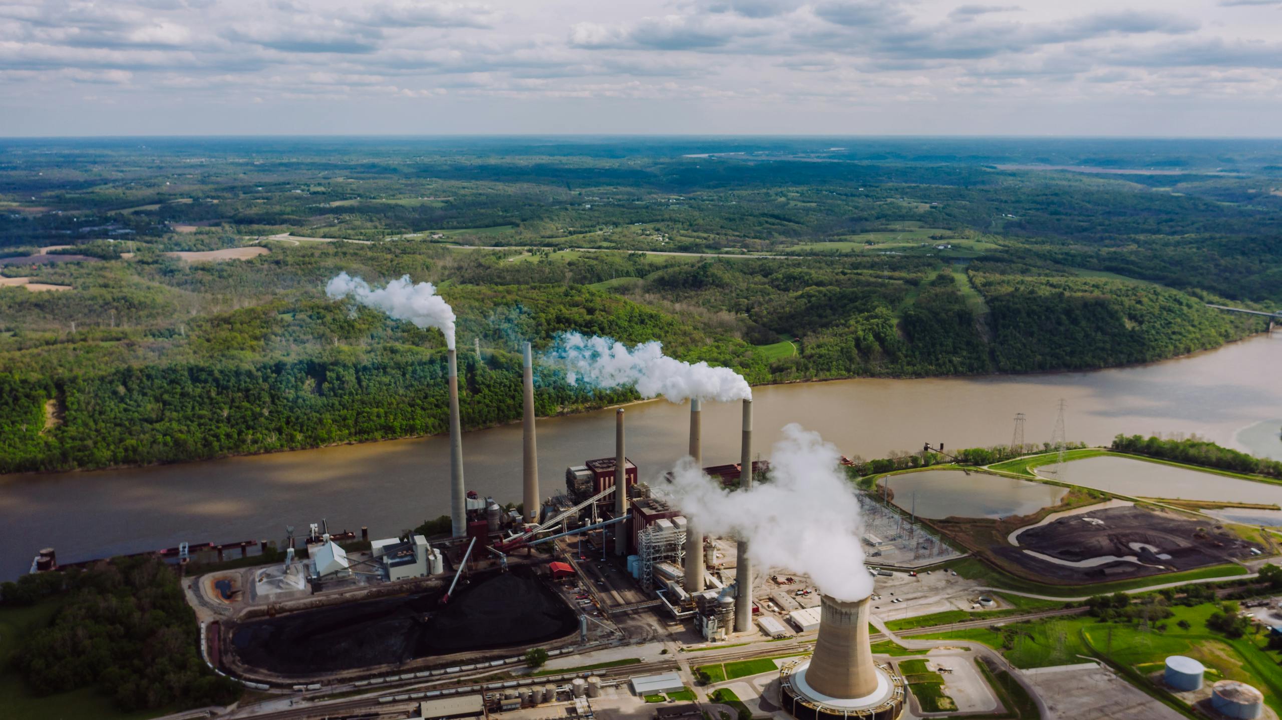Ohio Champion Trees Lewis Center Ohio Aerial view of an industrial plant with smokestacks emitting smoke by a river. Green landscape in the background.
