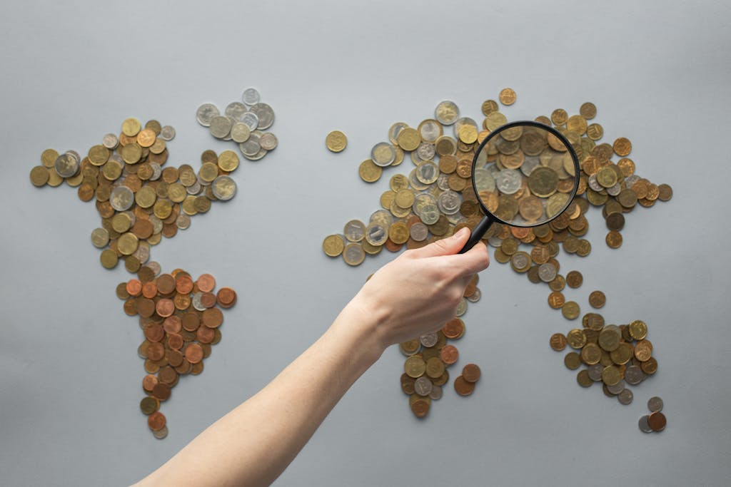 theglobeandmail.com.mx Top view of crop unrecognizable traveler with magnifying glass standing over world map made of various coins on gray background
