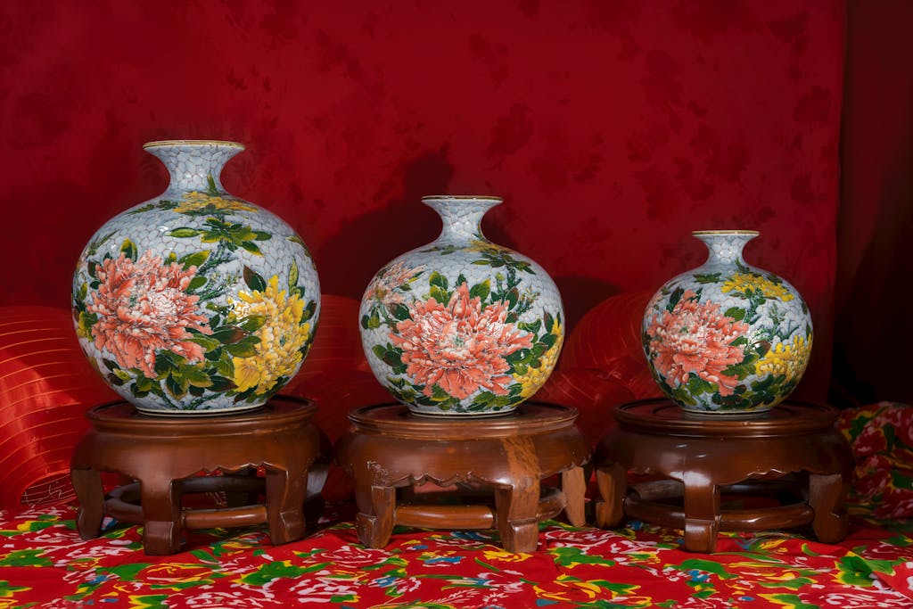 Shop Spring Vase Trio of artistic vases with colorful floral patterns on wooden stands, set against a rich red background.