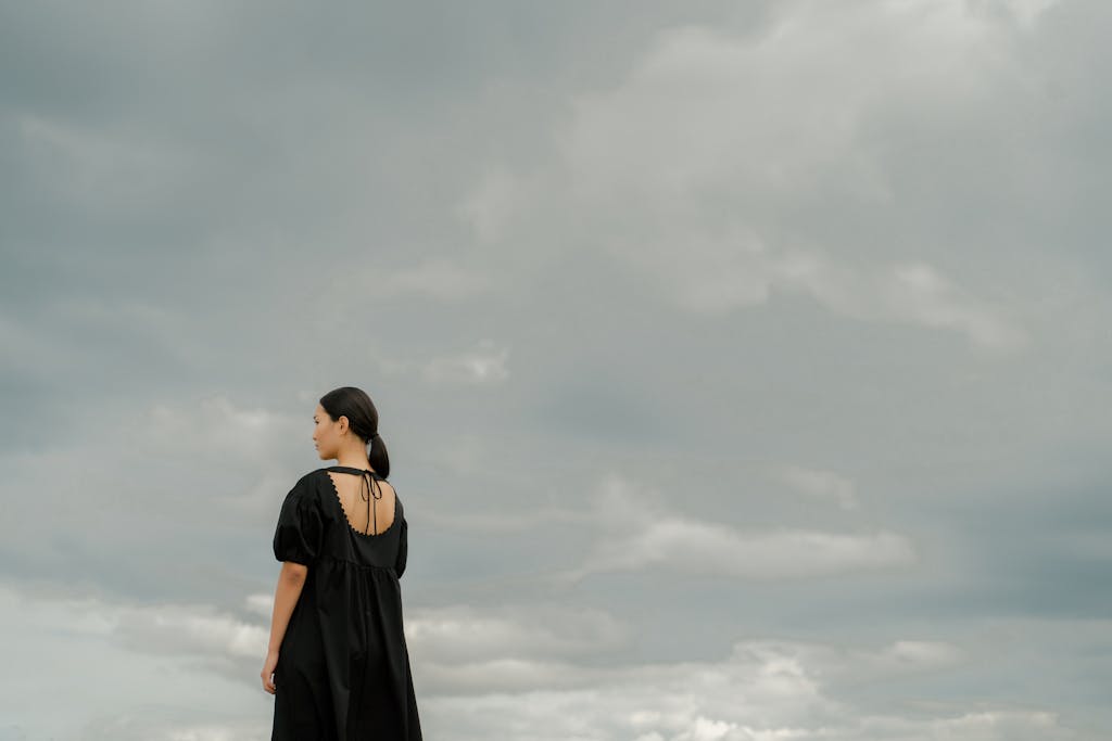 Carmela Clouth A woman in a black dress stands alone outdoors, gazing at a dramatic cloudy sky.