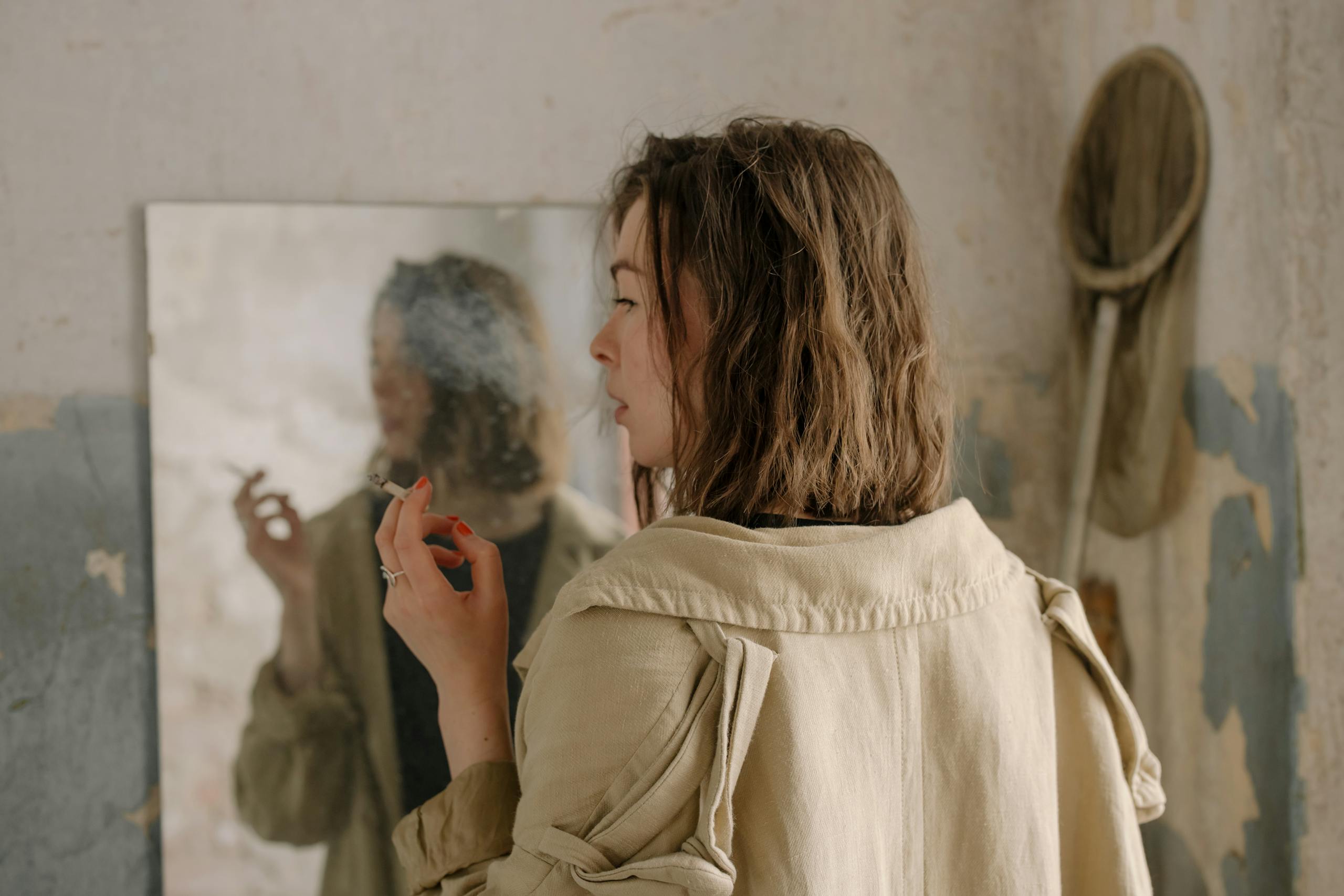 Carmela Clouth A woman in a trench coat smoking, reflected in a mirror against a shabby, textured wall.